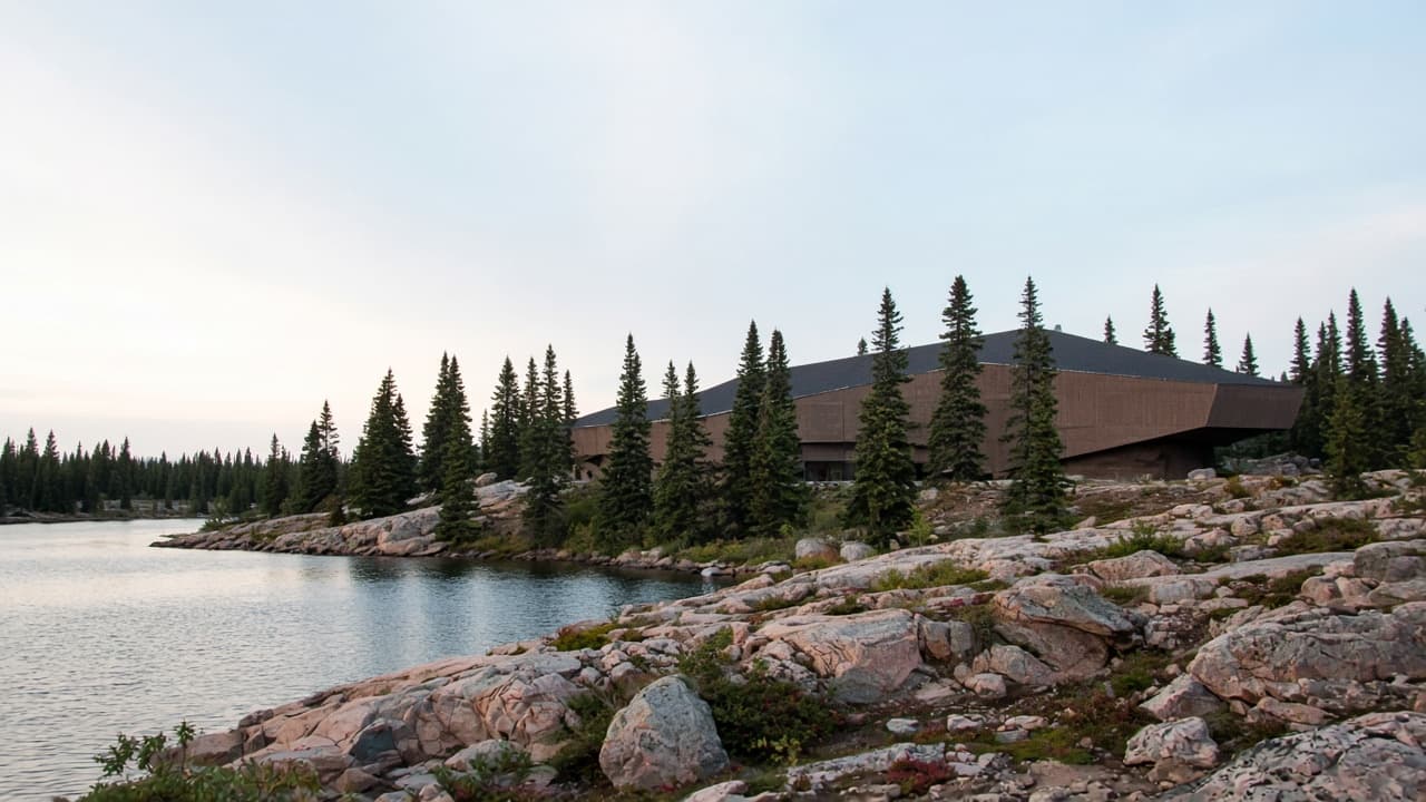 A low brown museum on Shield rock beside a lake, with spruce trees in front